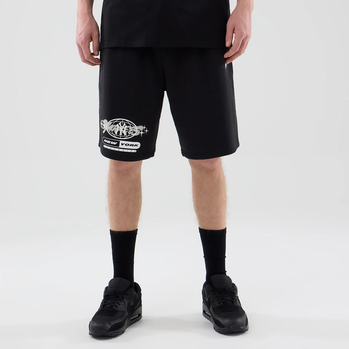 New Era New York Yankees MLB Logo Graphic Black Oversized Shorts, front view showing a printed Yankees logo in white and grey on the left leg, a loose fit with knee-length styling, worn with black trainers and socks for a casual look.