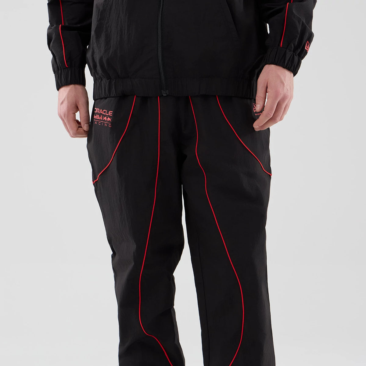 New Era Red Bull Racing Piping Track Black Track Pants, front view on a model showing red piping detail along the front seams, Oracle Red Bull Racing logo embroidered on the left thigh, and a relaxed fit with an elasticated waistband.
