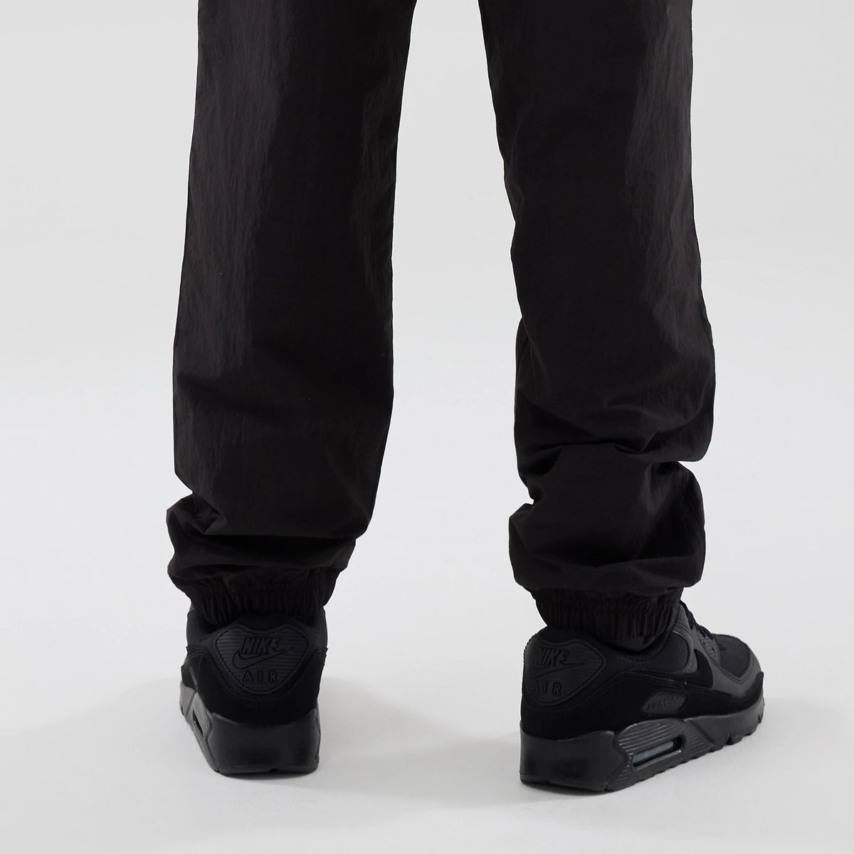 New Era Red Bull Racing Piping Track Black Track Pants, rear view on a model showing the relaxed fit with elasticated cuffs draping over black sneakers, highlighting the black fabric's texture and straight leg design.