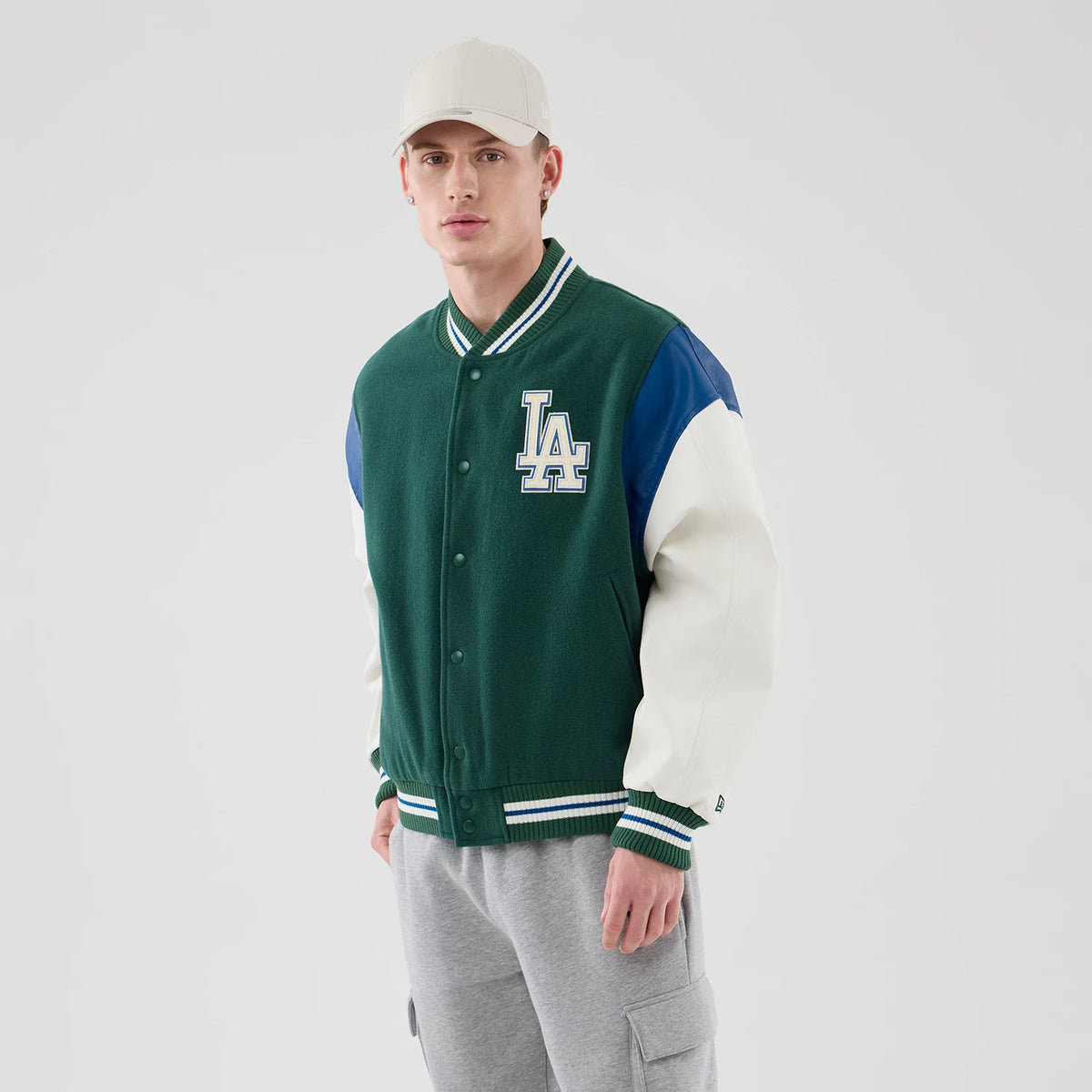 New Era LA Dodgers MLB Varsity Dark Green Jacket, front view on-model showing the white embroidered LA logo on the chest, dark green body with contrasting blue and white sleeves, and striped green and white ribbed collar, cuffs, and hem.