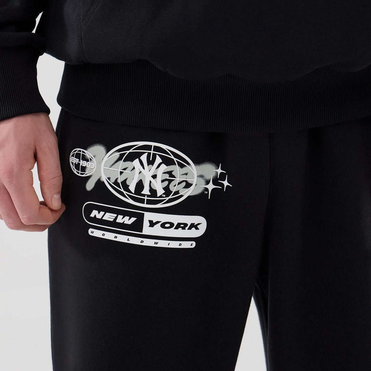 New Era New York Yankees MLB Logo Graphic Black Straight Leg Joggers, close-up showing the monochrome graphic featuring the New York Yankees logo in an oval, alongside New York and Worldwide lettering, printed on the left upper thigh.