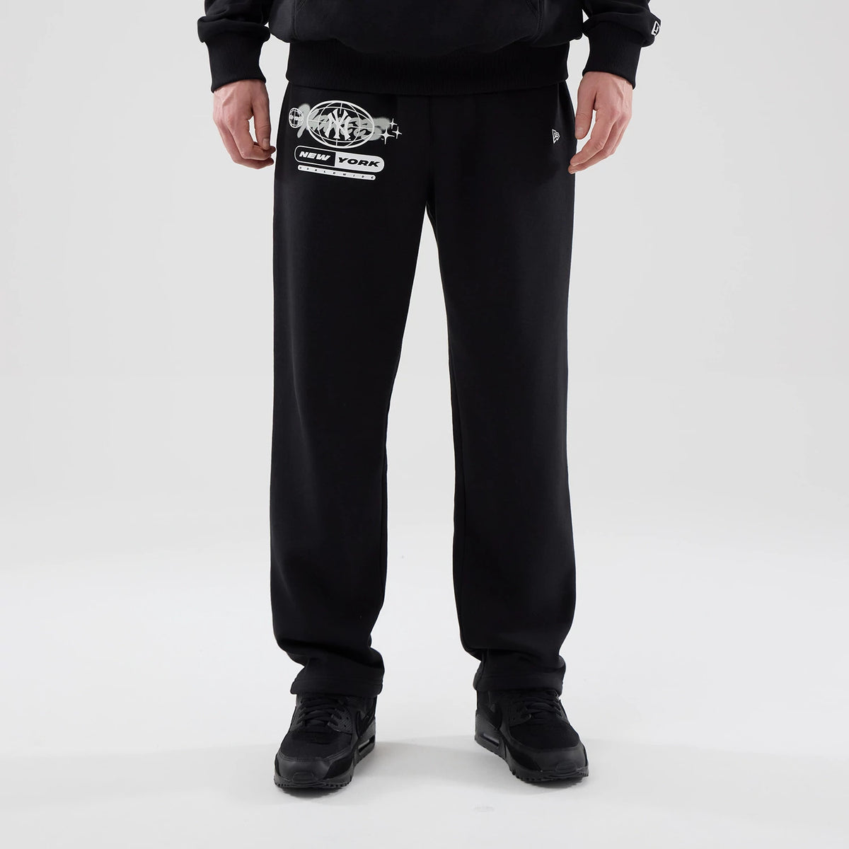 New Era New York Yankees MLB Logo Graphic Black Straight Leg Joggers, front view on a model wearing black trainers, featuring the New York Yankees logo with graphic details on the right thigh and the New Era flag logo on the left pocket.