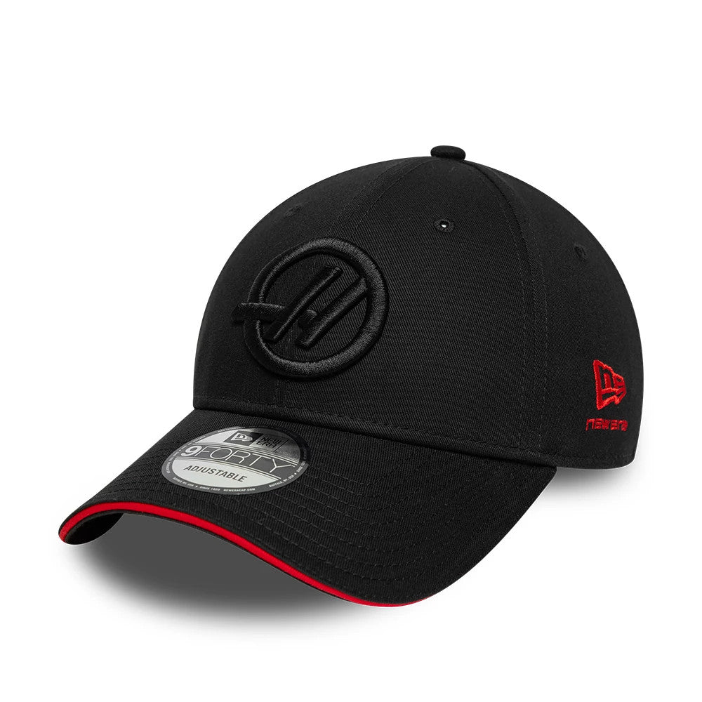 TGR Haas F1 Team Cotton Black New Era 9FORTY Adjustable Cap, three-quarter view showing an embroidered black Haas logo on the front panel, a red New Era flag logo on the left side, and a curved visor with a red edge and a 9FORTY size sticker.