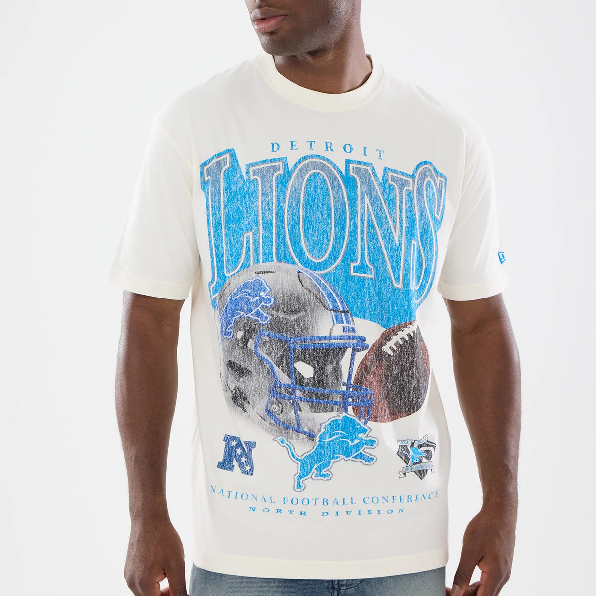 New Era Detroit Lions NFL Sport Classic Cream T-Shirt, front view on a model showing large distressed blue LIONS lettering across the chest, a detailed football helmet graphic with a blue lion logo, and small embroidered New Era logo on the sleeve.