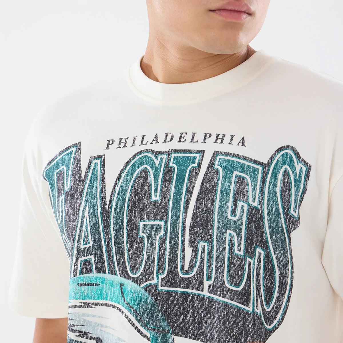 New Era Philadelphia Eagles NFL Sport Classic Cream T-Shirt, front view on a model showing a large distressed Eagles graphic across the chest in green and black with Philadelphia text above, under a rounded cream neckline and short sleeves.