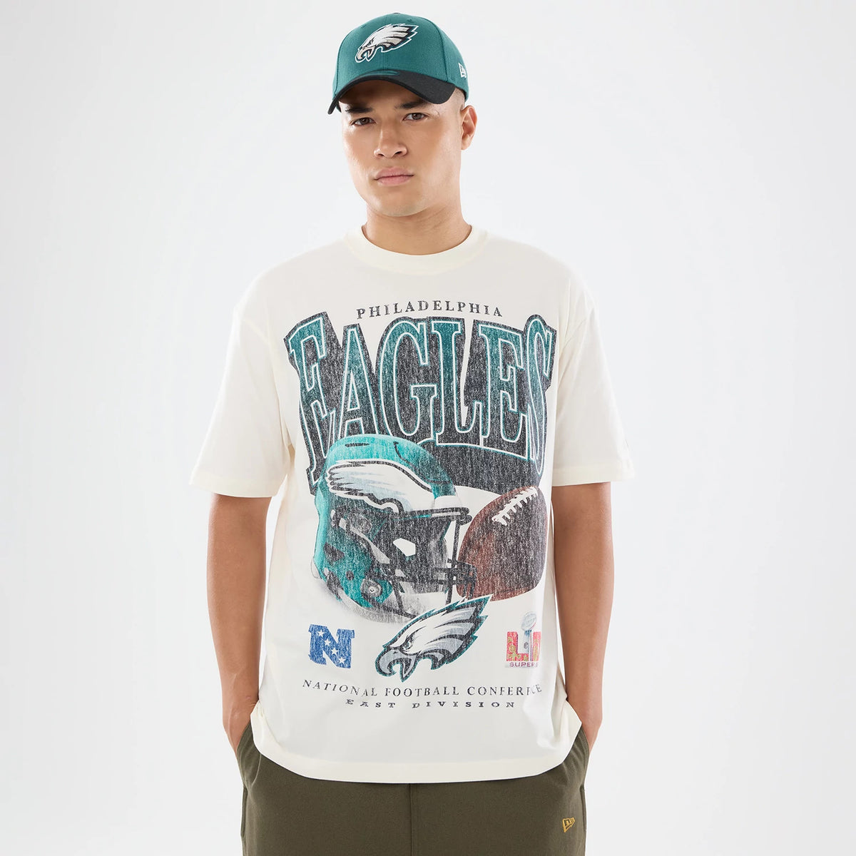 New Era Philadelphia Eagles NFL Sport Classic Cream T-Shirt, front view on-model displaying the large printed Eagles helmet graphic and lettering across the chest, styled with a matching green cap featuring the embroidered Eagles logo.