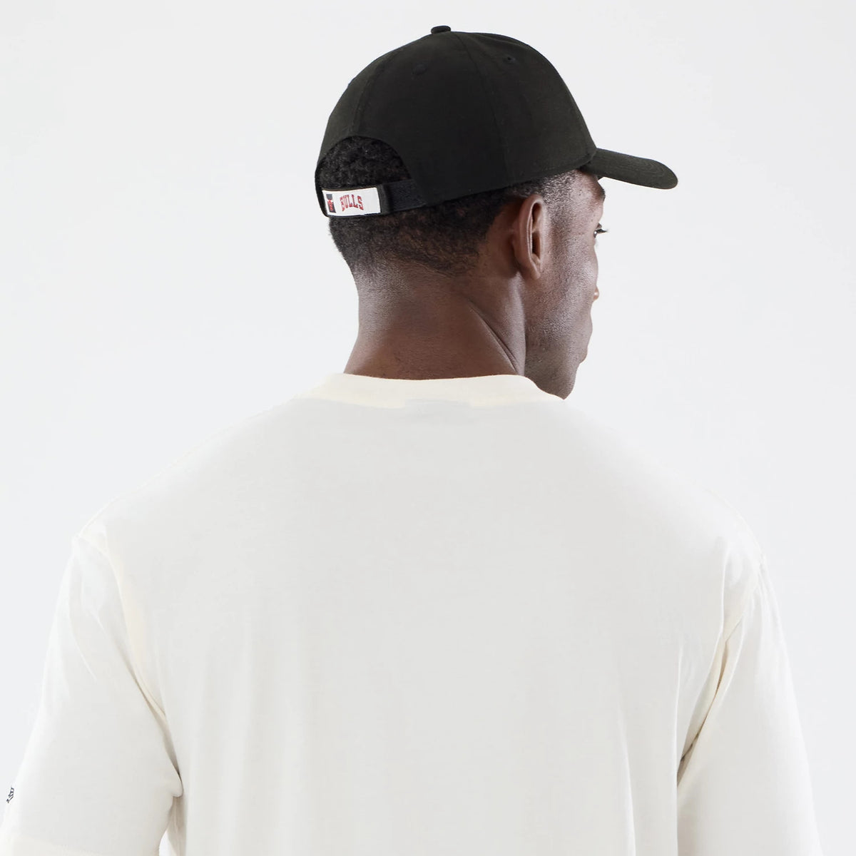 New Era Chicago Bulls NBA Sport Classic Cream T-Shirt, back view on a model wearing a dark cap, highlighting the plain cream fabric of the shirt with a relaxed fit, and a small Bulls logo label on the back strap of the cap.
