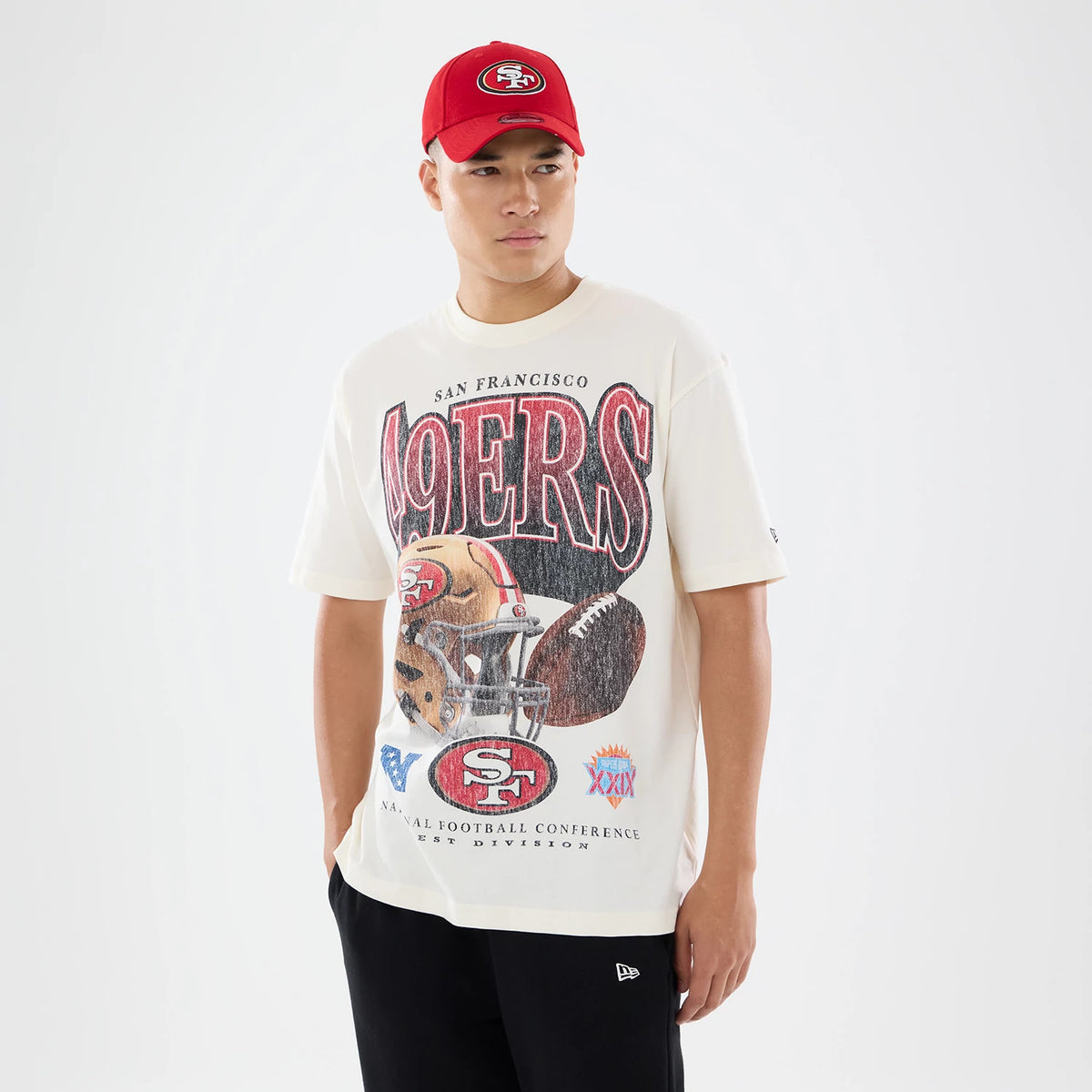 New Era San Francisco 49ers NFL Sport Classic Cream T-Shirt, front view on-model showing large 49ERS graphic with a helmet and football, SAN FRANCISCO lettering above, and NFL motifs below, the model styled with a red cap and black trousers.