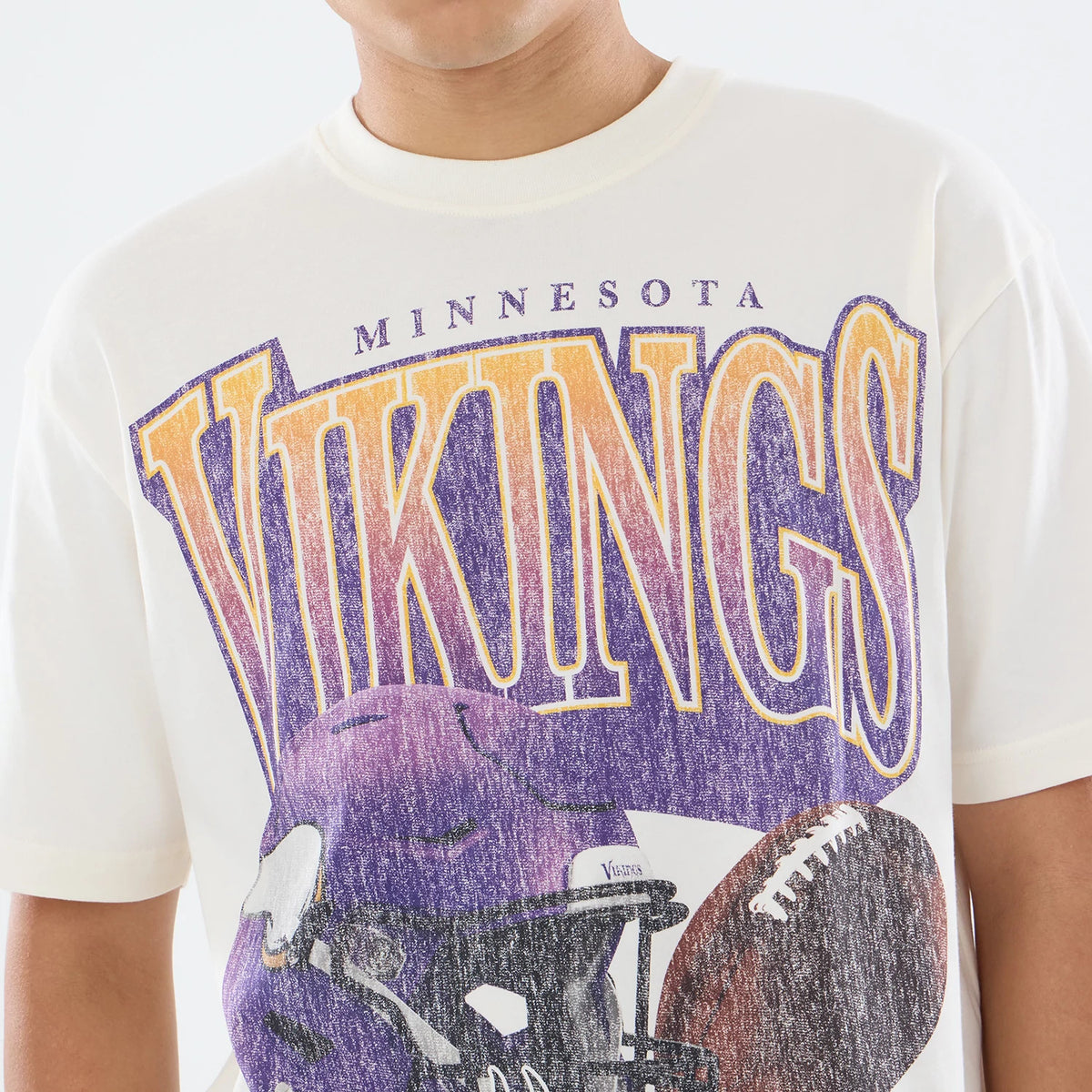 New Era Minnesota Vikings NFL Sport Classic Cream T-Shirt, front view featuring the oversized Vikings lettering in purple and gold across the chest, a faded purple football helmet graphic below, and portions of a football.