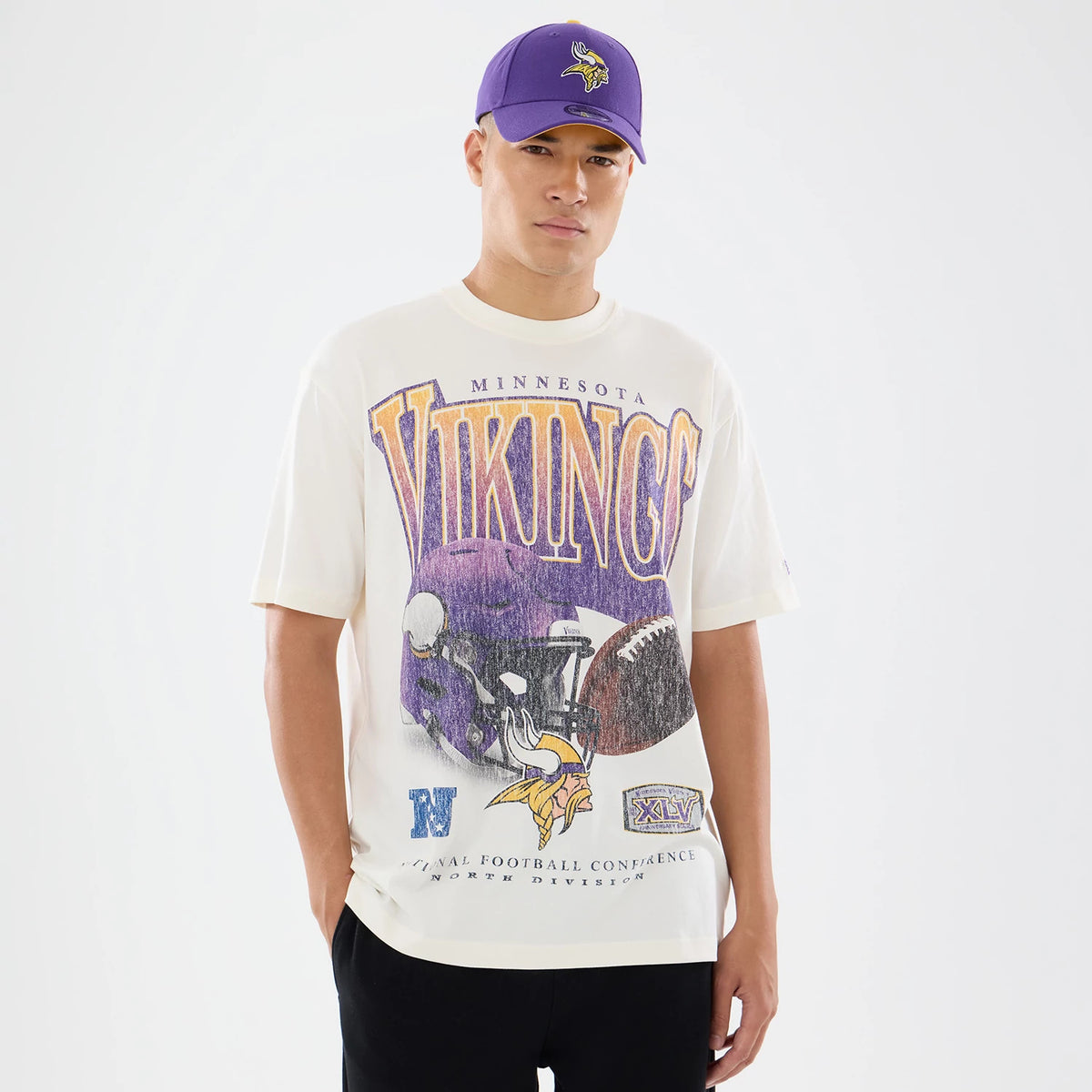New Era Minnesota Vikings NFL Sport Classic Cream T-Shirt, front view on a model with a purple cap showing a large purple and yellow Vikings graphic across the chest, featuring a helmet and football imagery below lettering.