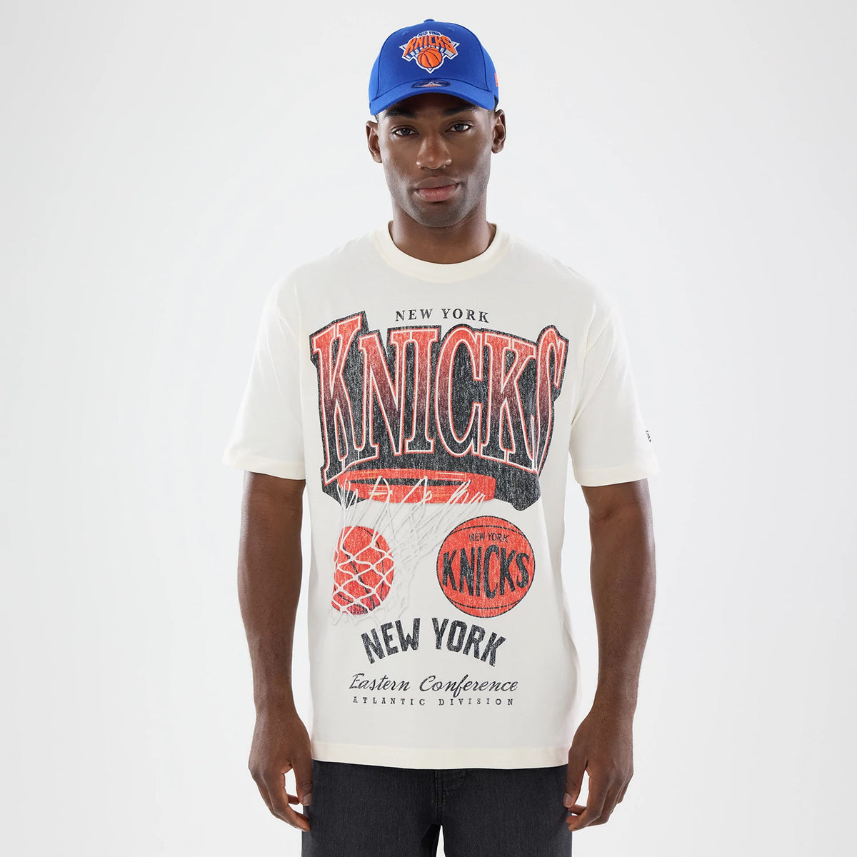 New Era New York Knicks NBA Sport Classic Cream T-Shirt, front view on a model, displaying black and red KNICKS lettering across the chest with a basketball hoop graphic underneath, and a small New Era logo on the left sleeve.