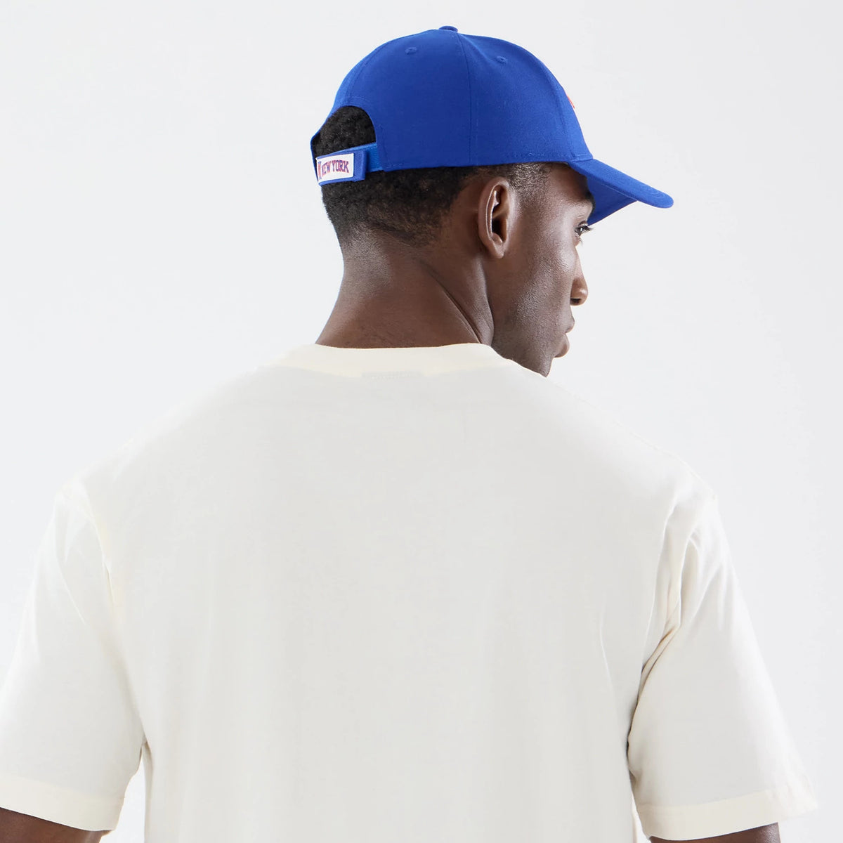 New Era New York Knicks NBA Sport Classic Cream T-Shirt, back view on a model with a blue cap, showing a plain cream back with short sleeves and a ribbed neckline, complemented by the contrast of the cap's New York lettering on the rear strap.