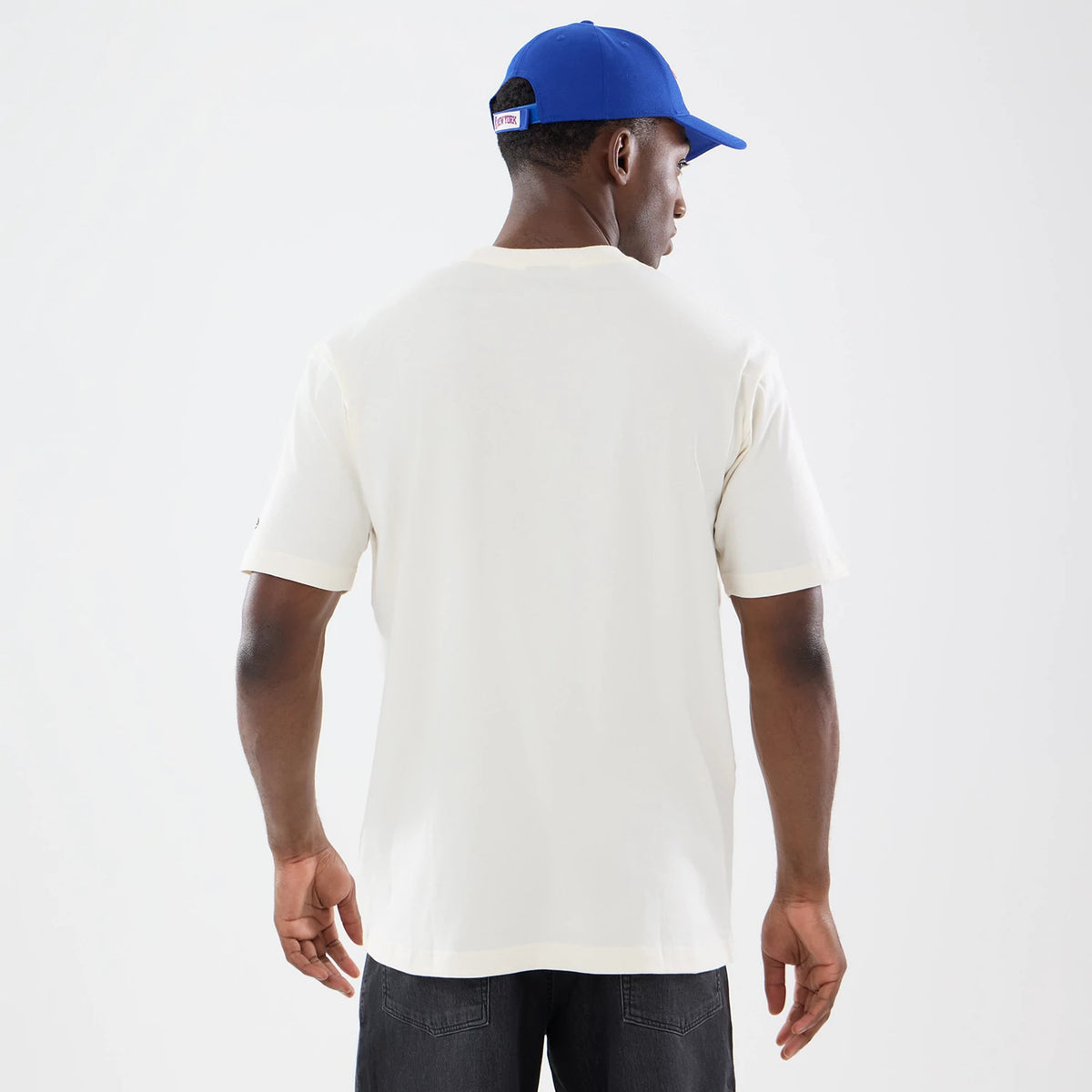 New Era New York Knicks NBA Sport Classic Cream T-Shirt, back view on a model with a blue cap, showing the plain cream back and short sleeves, with a small black embroidered logo on the left sleeve near the cuff.