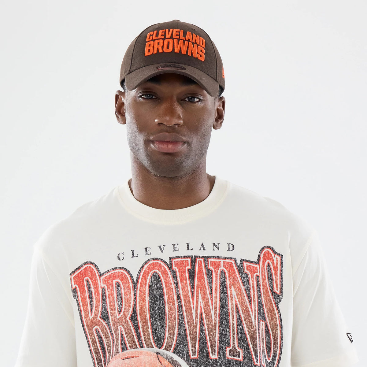 New Era Cleveland Browns NFL Sport Classic Cream T-Shirt, front view on a model wearing a brown cap with orange embroidered Cleveland Browns text, the cream T-shirt features large distressed Browns lettering and a football graphic across the chest.