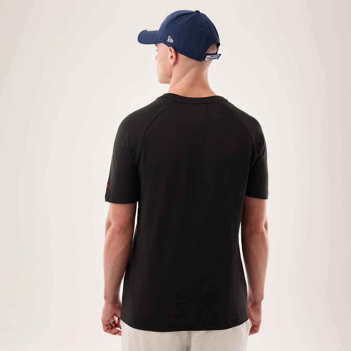New Era New England Patriots NFL Sideline 2026 Black T-Shirt, back view on a model, showing a plain black back with short sleeves, a small red embroidered Patriots logo on the right sleeve and paired with a blue cap.