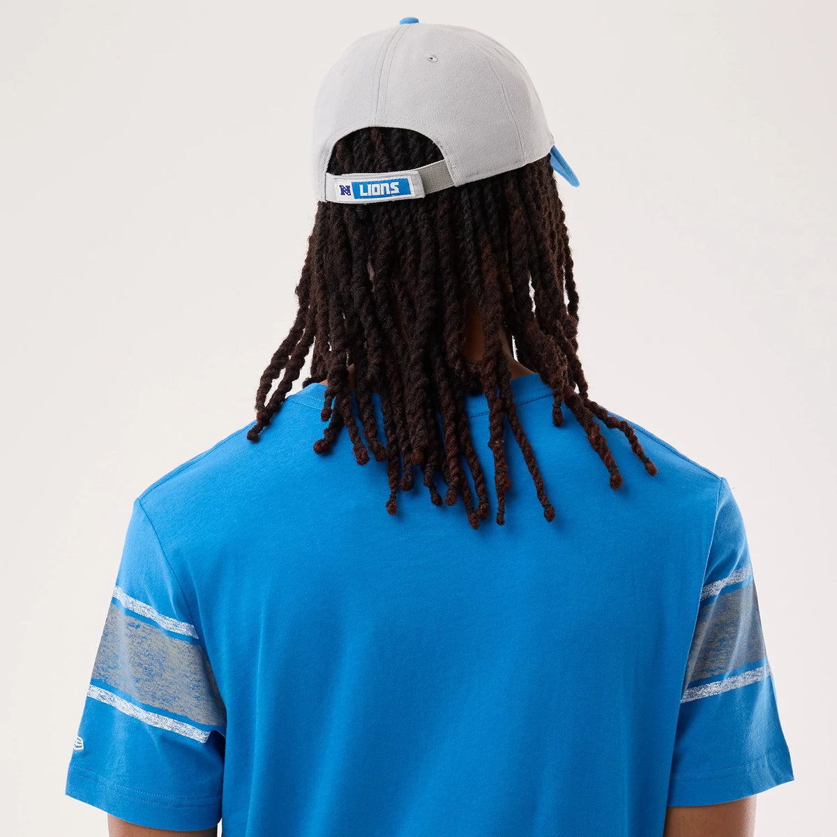 New Era Detroit Lions NFL Sideline 2026 Blue T-Shirt, back view on a model with shoulder-length hair, featuring grey and white patterned stripes on the sleeves, and a close-up of the blue fabric texture.