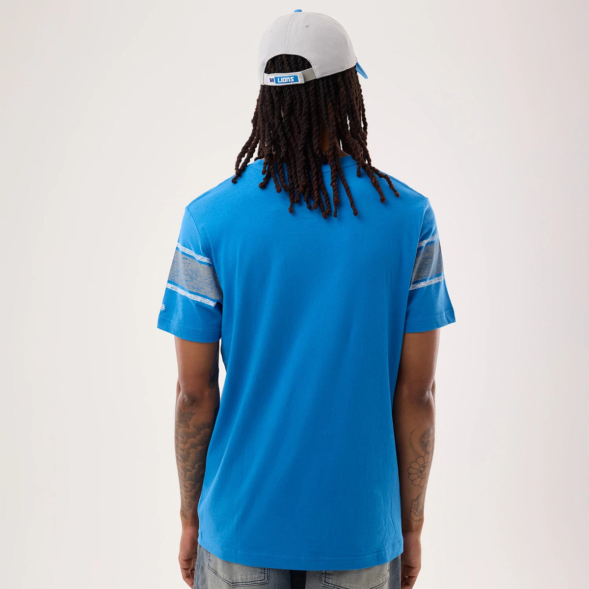 New Era Detroit Lions NFL Sideline 2026 Blue T-Shirt, back view on a model wearing a cap, showing the blue colour with grey and blue stripes on the sleeves, and an embroidered New Era logo on the right sleeve near the hem.