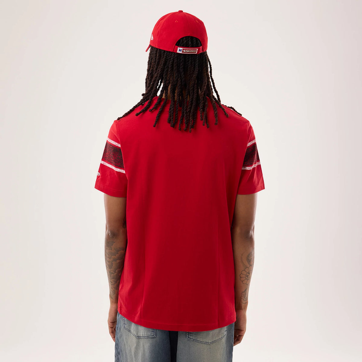 New Era San Francisco 49ers NFL Sideline 2026 Red T-Shirt, back view on a model with a matching red cap, displaying plain red fabric across the back, horizontal striped on the sleeves, and the New Era logo embroidered on the left sleeve.