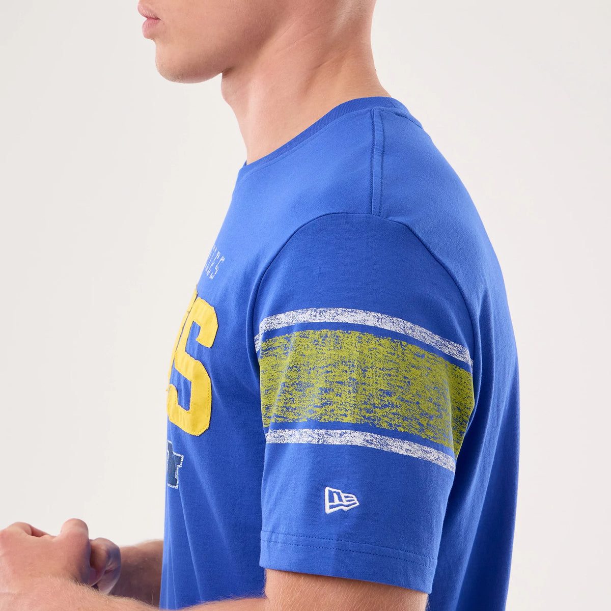 New Era LA Rams NFL Sideline 2026 Bright Blue T-Shirt, side view on a model showing a distressed yellow and white stripe on the sleeve, large yellow graphic text across the chest, and a white embroidered New Era logo above the hem on the sleeve.