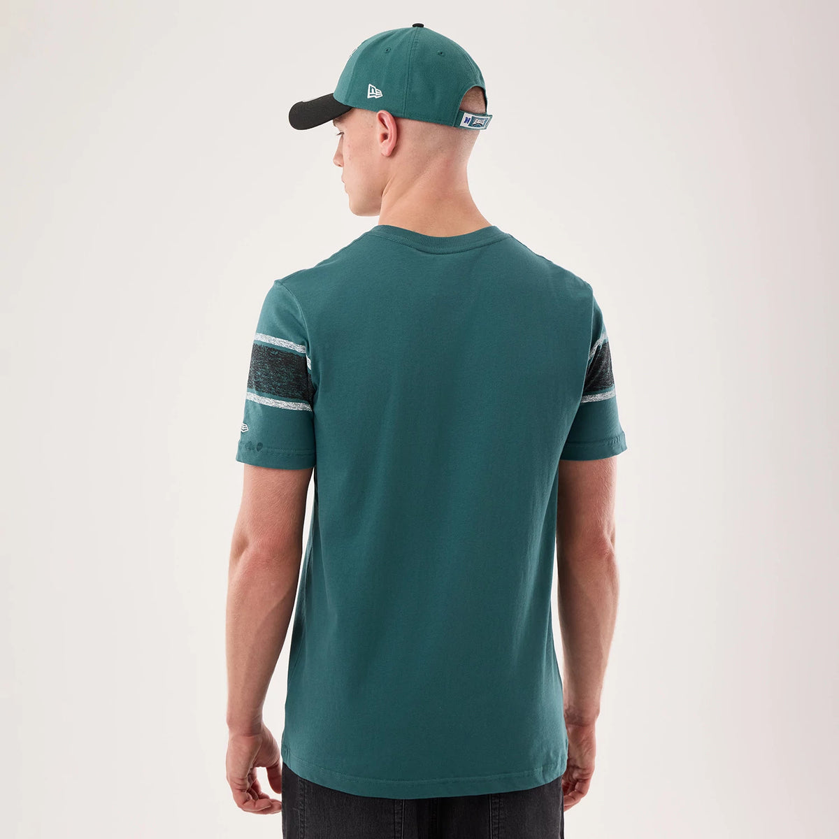 New Era Philadelphia Eagles NFL Sideline 2026 Dark Green T-Shirt, back view on a model wearing a matching cap, showing the plain dark green back with striped details on the sleeves and a small raised New Era logo near the left sleeve edge.