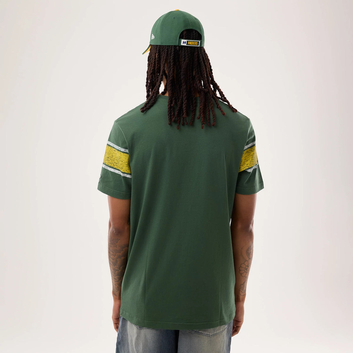 New Era Green Bay Packers NFL Sideline 2026 Dark Green T-Shirt, back view on a model wearing a matching cap, showing two yellow stripe bands with a distressed effect on each sleeve and a plain dark green back without additional graphics.