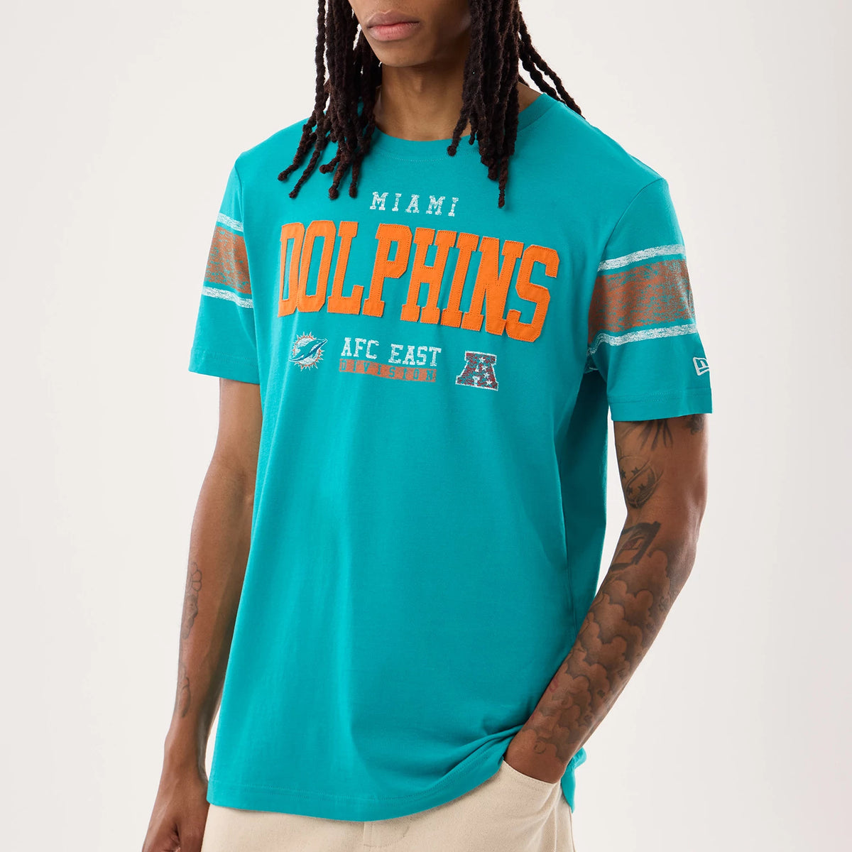 New Era Miami Dolphins NFL Sideline 2026 Turquoise T-Shirt, front view on a model showing large orange Dolphins lettering across the chest, a team logo and AFC East graphic below, and striped on the sleeves.