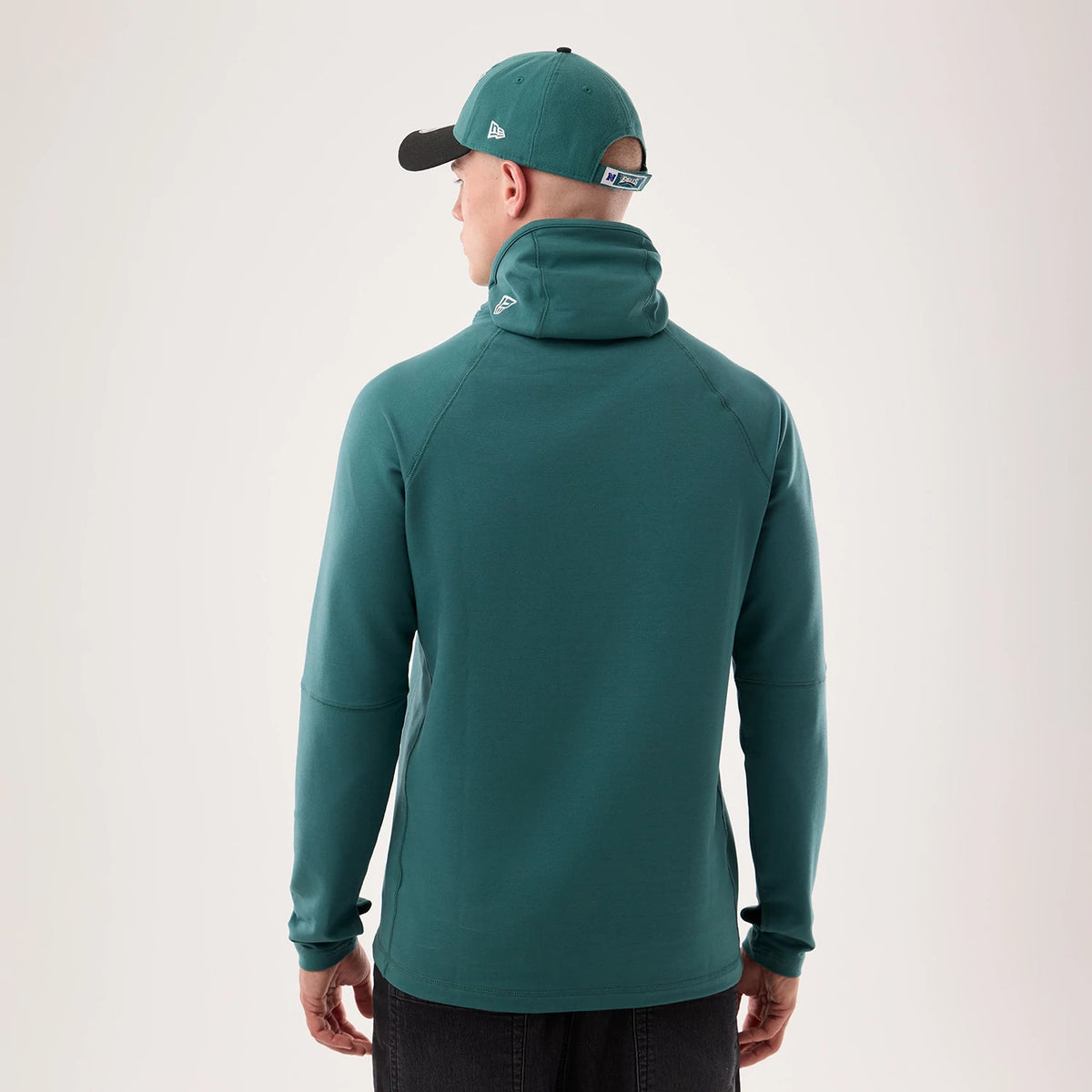 New Era Philadelphia Eagles NFL Sideline 2026 Dark Green Balaclava Hoodie, back view on a model wearing a matching dark green cap with a black visor, showing the raised New Era flag logo on the high collar and another logo on the cap.