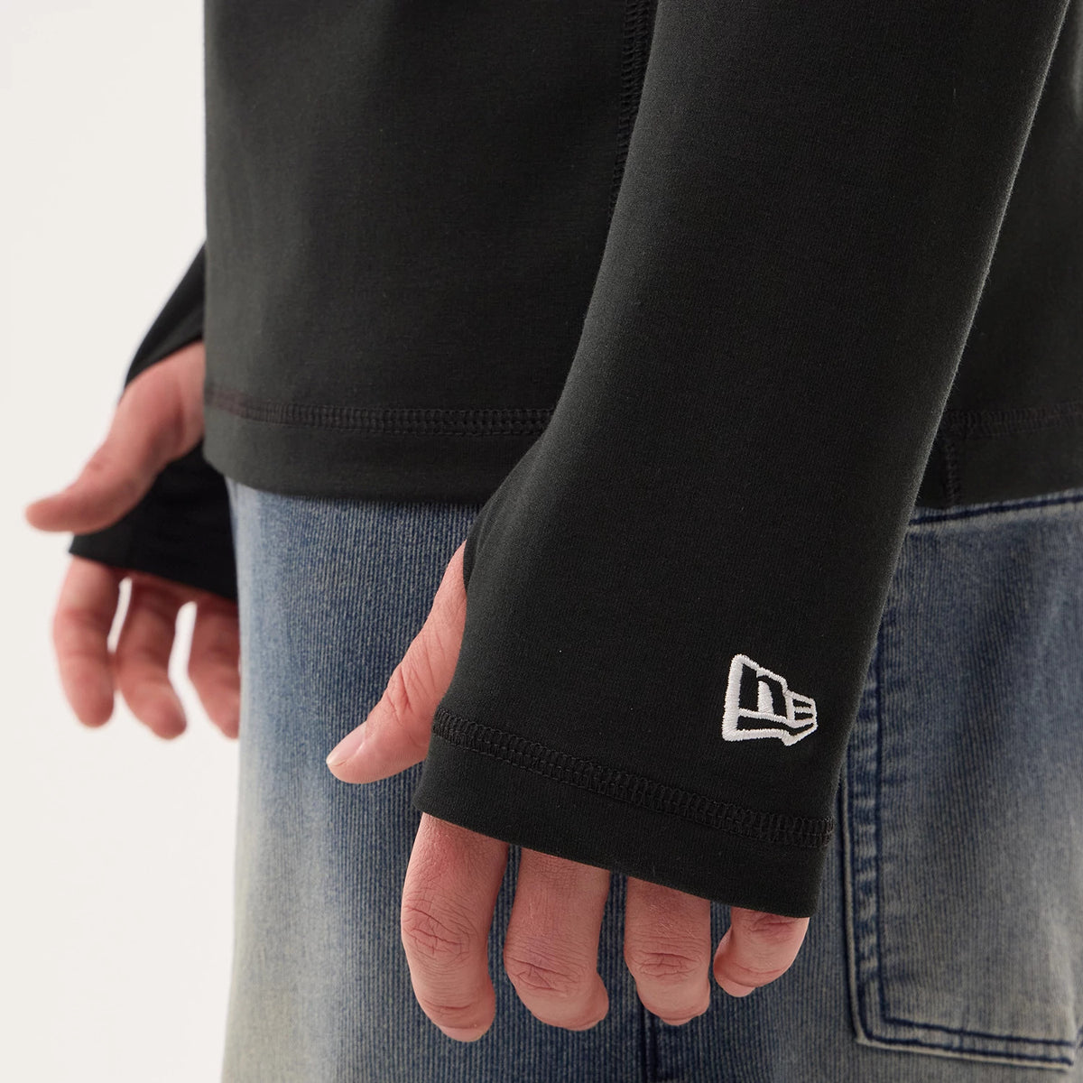 New Era Las Vegas Raiders NFL Sideline 2026 Black Balaclava Hoodie, on-model view showing the sleeve with a thumbhole design, an embroidered white New Era logo near the wrist, and styled with faded blue denim jeans.