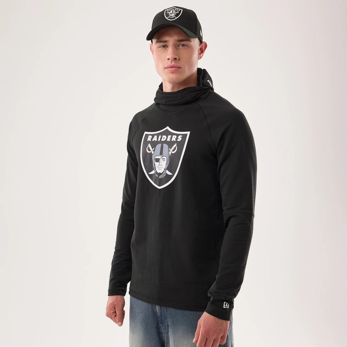 New Era Las Vegas Raiders NFL Sideline 2026 Black Balaclava Hoodie, front view on a model styled with a matching Raiders cap, showing the prominent embroidered Raiders shield logo on the chest.