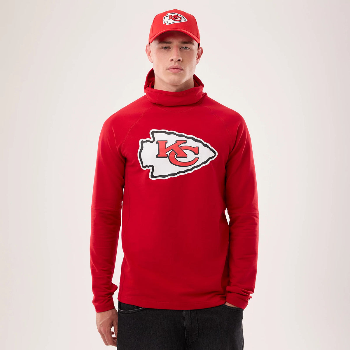 New Era Kansas City Chiefs NFL Sideline 2026 Red Balaclava Hoodie, front view on a model with a matching cap, featuring a large embroidered Chiefs logo across the chest, a unique balaclava-style neckline, and long sleeves extending to the wrists.