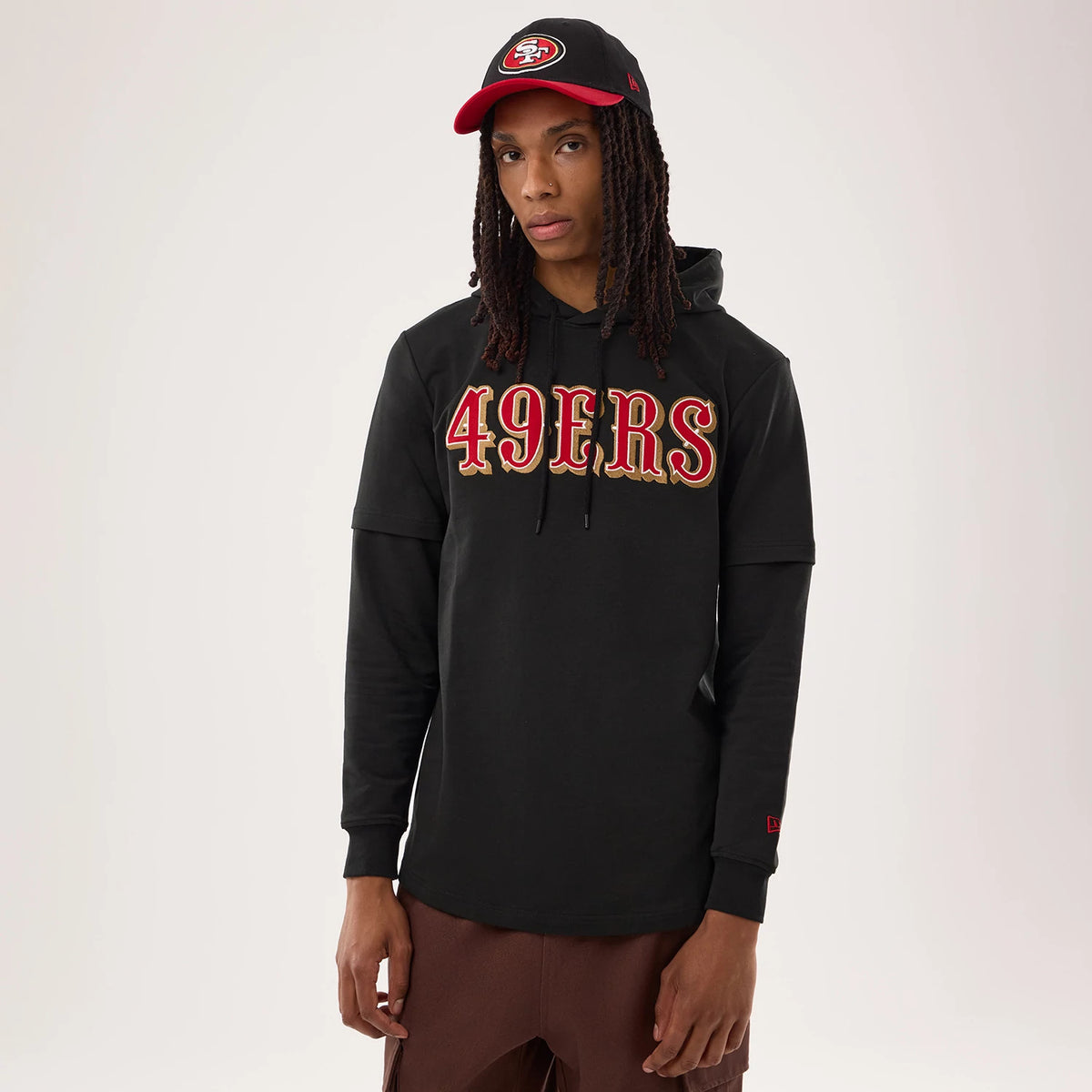 New Era San Francisco 49ers NFL Sideline 2026 Black Double Sleeve Hoodie, front view on a model wearing a matching cap, featuring large embroidered 49ERS lettering in red and gold across the chest and a New Era logo on the left cuff.