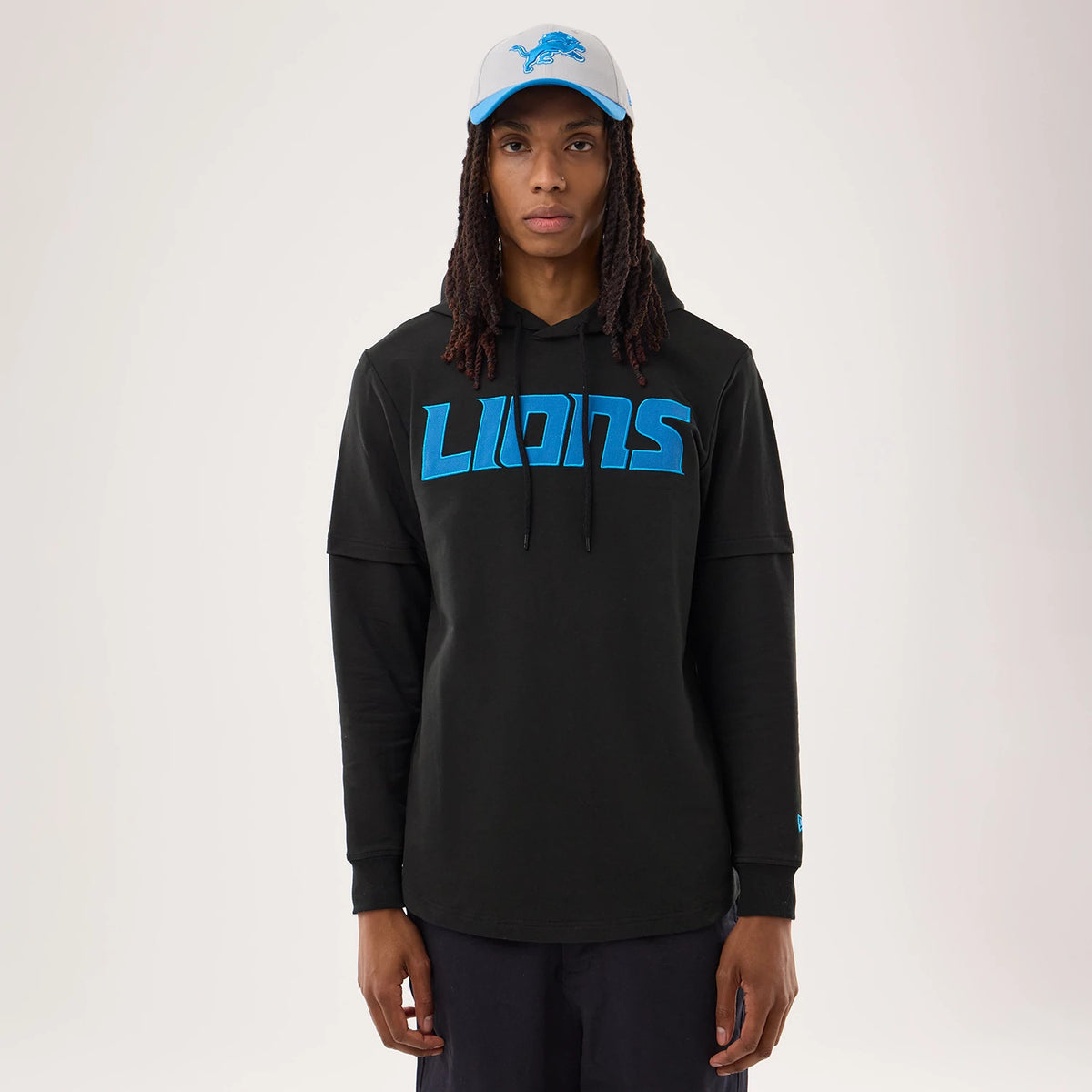 New Era Detroit Lions NFL Sideline 2026 Black Double Sleeve Hoodie, front view on a model styled with a contrasting cap, featuring blue LIONS lettering across the chest, double-layered sleeves and an embroidered New Era logo on the left wrist.