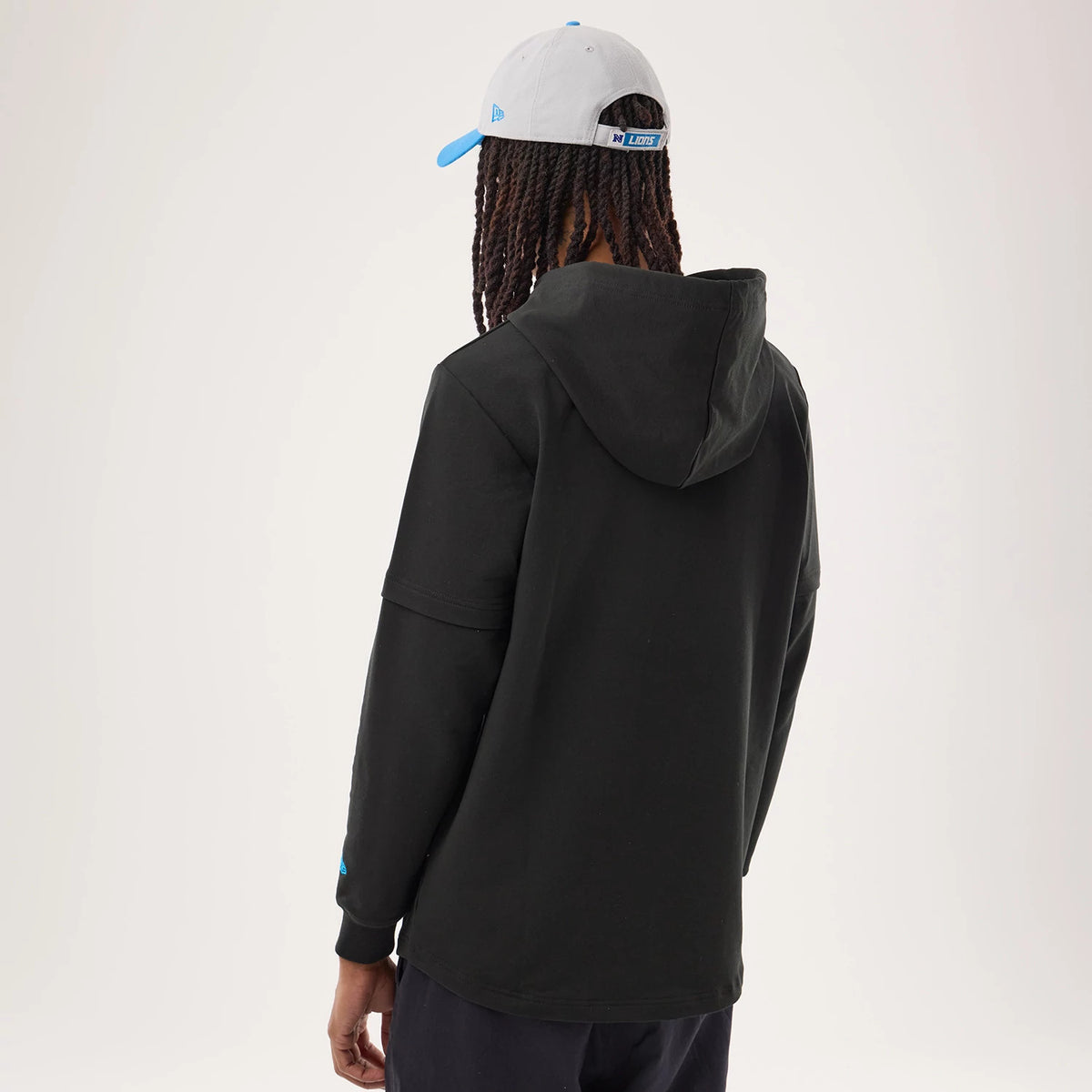 New Era Detroit Lions NFL Sideline 2026 Black Double Sleeve Hoodie, back view on a model styled with a grey cap featuring a blue visor, showing the simple black hood, dropped shoulder seams, and a small raised New Era logo on the left sleeve.