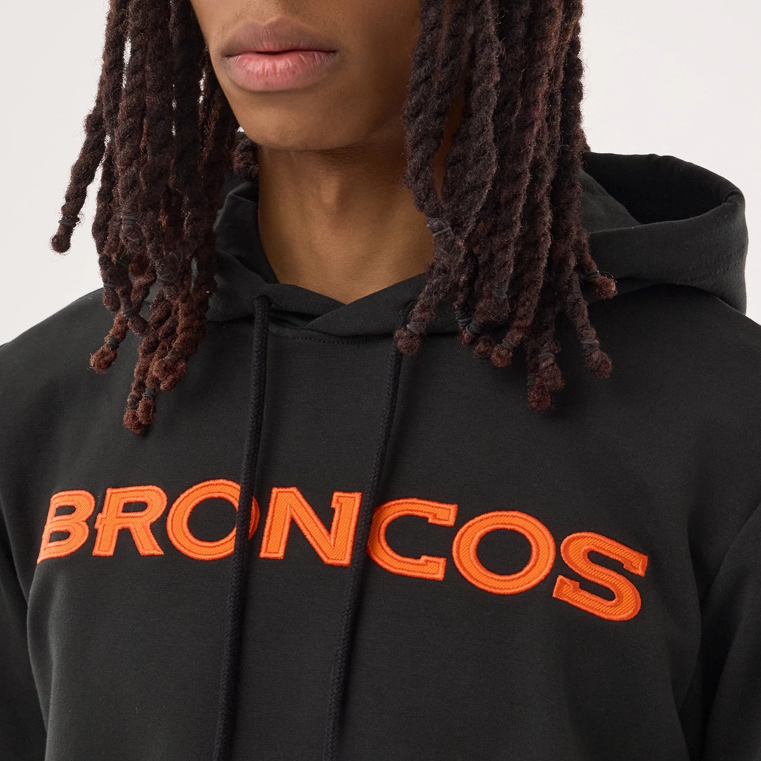New Era Denver Broncos NFL Sideline 2026 Black Double Sleeve Hoodie, front view on-model showing an orange embroidered BRONCOS logo across the chest, black drawstrings from the hood, and a black fabric finish, styled with shoulder-length locks.