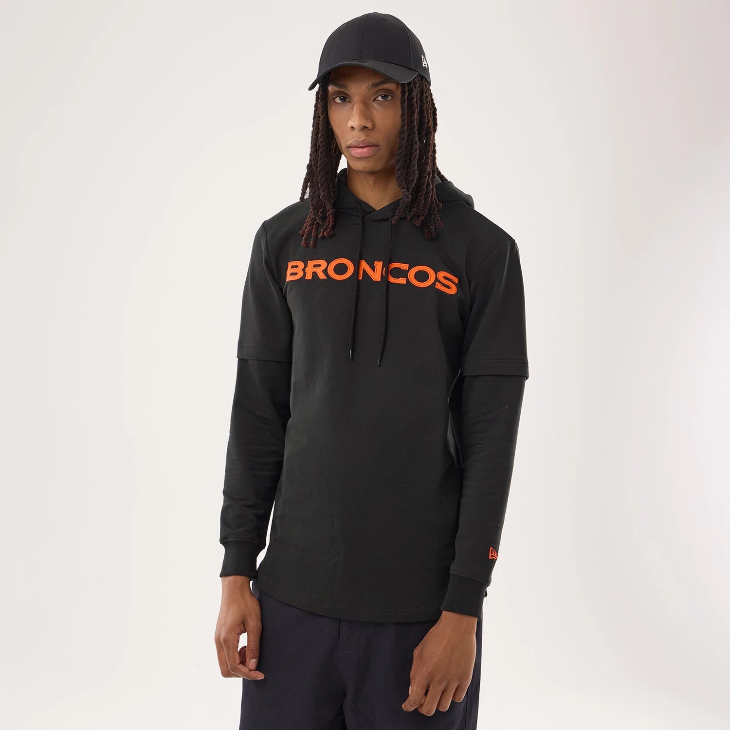 New Era Denver Broncos NFL Sideline 2026 Black Double Sleeve Hoodie, front view on-model showing the orange Broncos lettering across the chest, layered black sleeves with ribbed cuffs, and an embroidered New Era logo on the left sleeve.