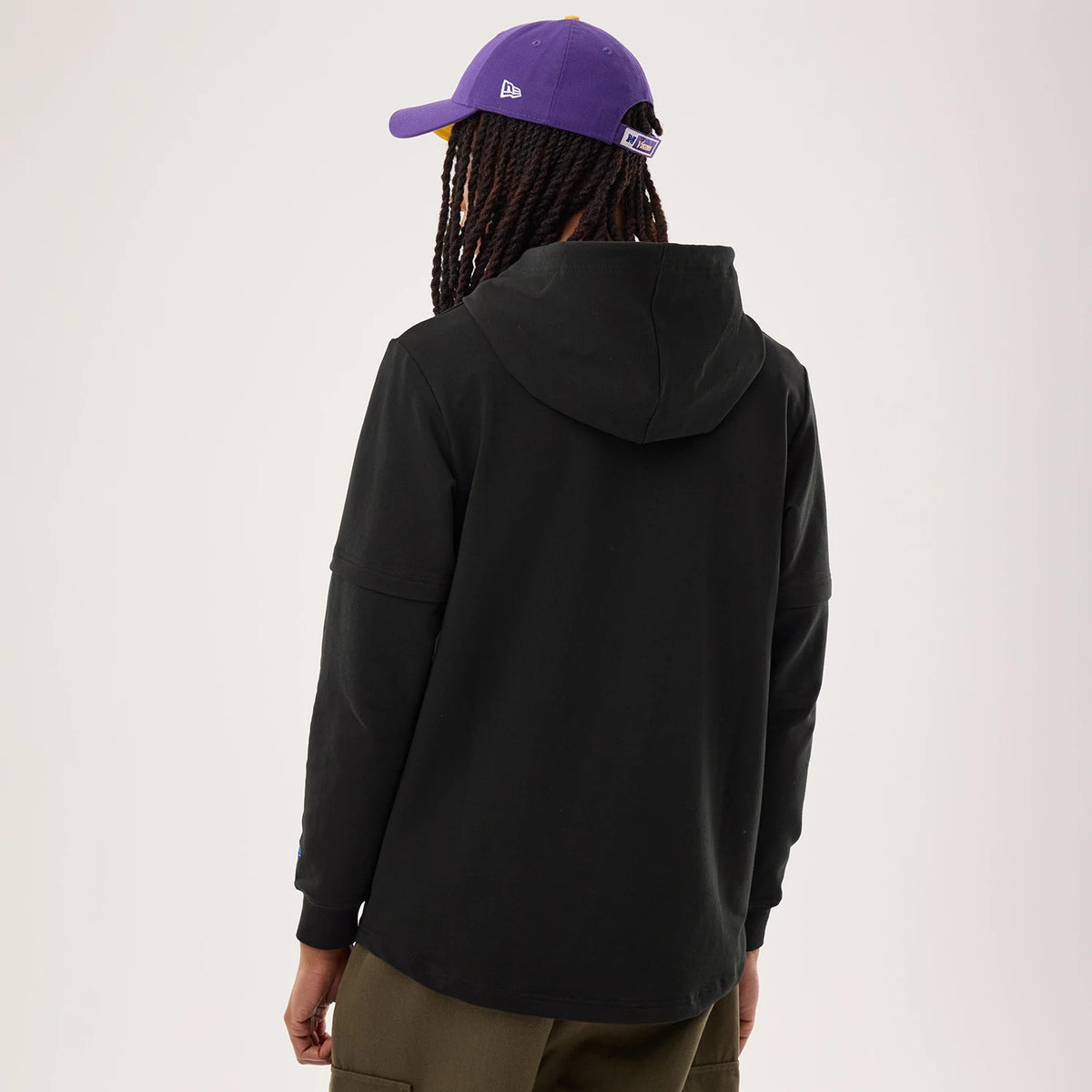 New Era Minnesota Vikings NFL Sideline 2026 Black Double Sleeve Hoodie, back view on a model wearing a purple cap showing the simple black hood, seam detail on dropped shoulders and relaxed fit extending to elongated sleeves with cuff design.