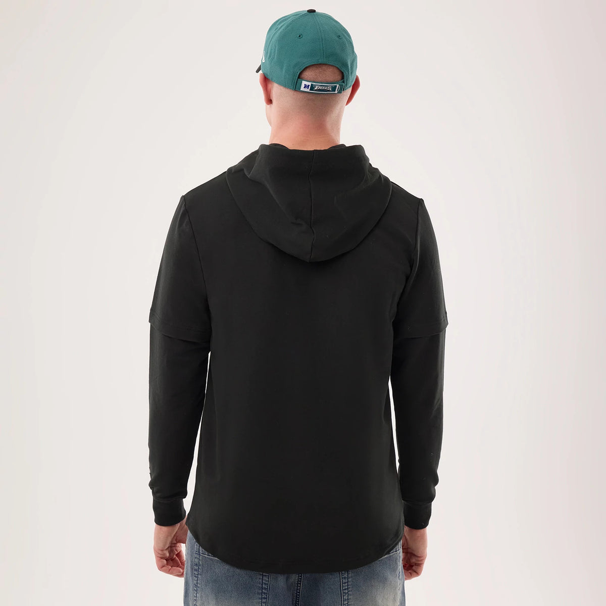 New Era Philadelphia Eagles NFL Sideline 2026 Black Double Sleeve Hoodie, back view on a model wearing a teal cap, showing the attached hood draped down the back, long black sleeves, and a slightly curved hemline above blue jeans.