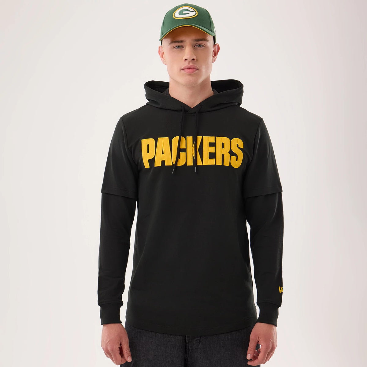 New Era Green Bay Packers NFL Sideline 2026 Black Double Sleeve Hoodie, front view on a model also wearing a cap, featuring yellow PACKERS lettering across the chest, long sleeves with double layering, and an embroidered New Era logo on the sleeve.
