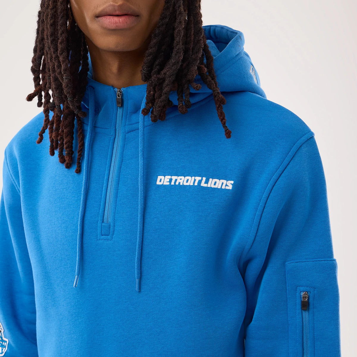 New Era Detroit Lions NFL Sideline 2026 Blue Pullover Hoodie, front view on a model showing the white embroidered Detroit Lions logo on the left chest, a short zip under the neckline, and a small zipped pocket on the right sleeve.