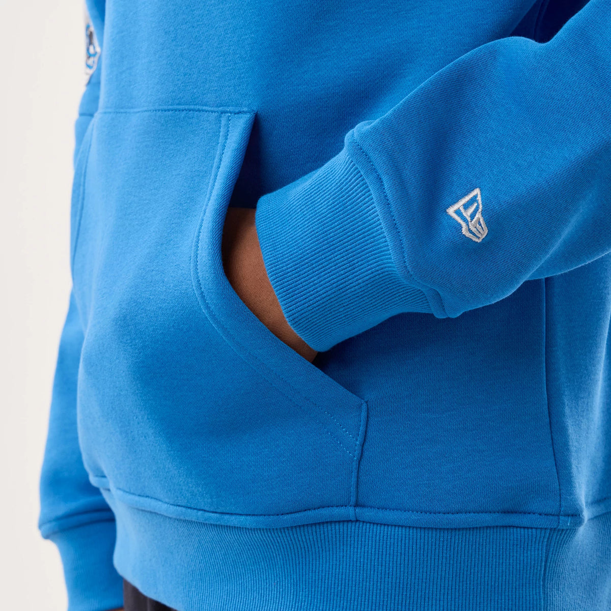New Era Detroit Lions NFL Sideline 2026 Blue Pullover Hoodie, front view showing a close-up of the kangaroo pocket with a hand partially inside, embroidered New Era logo on the cuff, and a blue colour covering the whole garment.