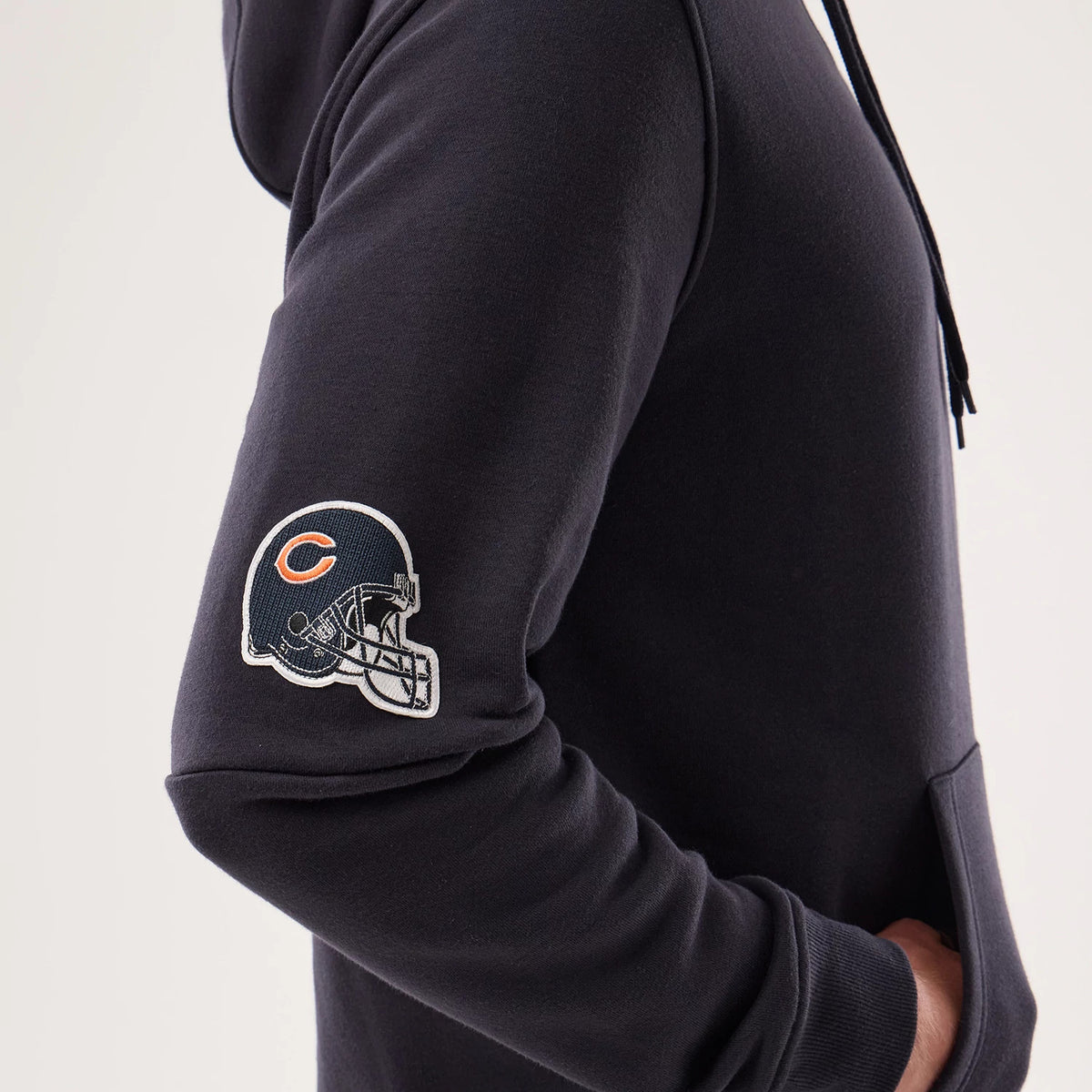 New Era Chicago Bears NFL Sideline 2026 Navy Pullover Hoodie, side view showcasing an embroidered Chicago Bears helmet logo on the sleeve, a relaxed fit with ribbed cuffs, and a model styling it with one hand in the front pocket.