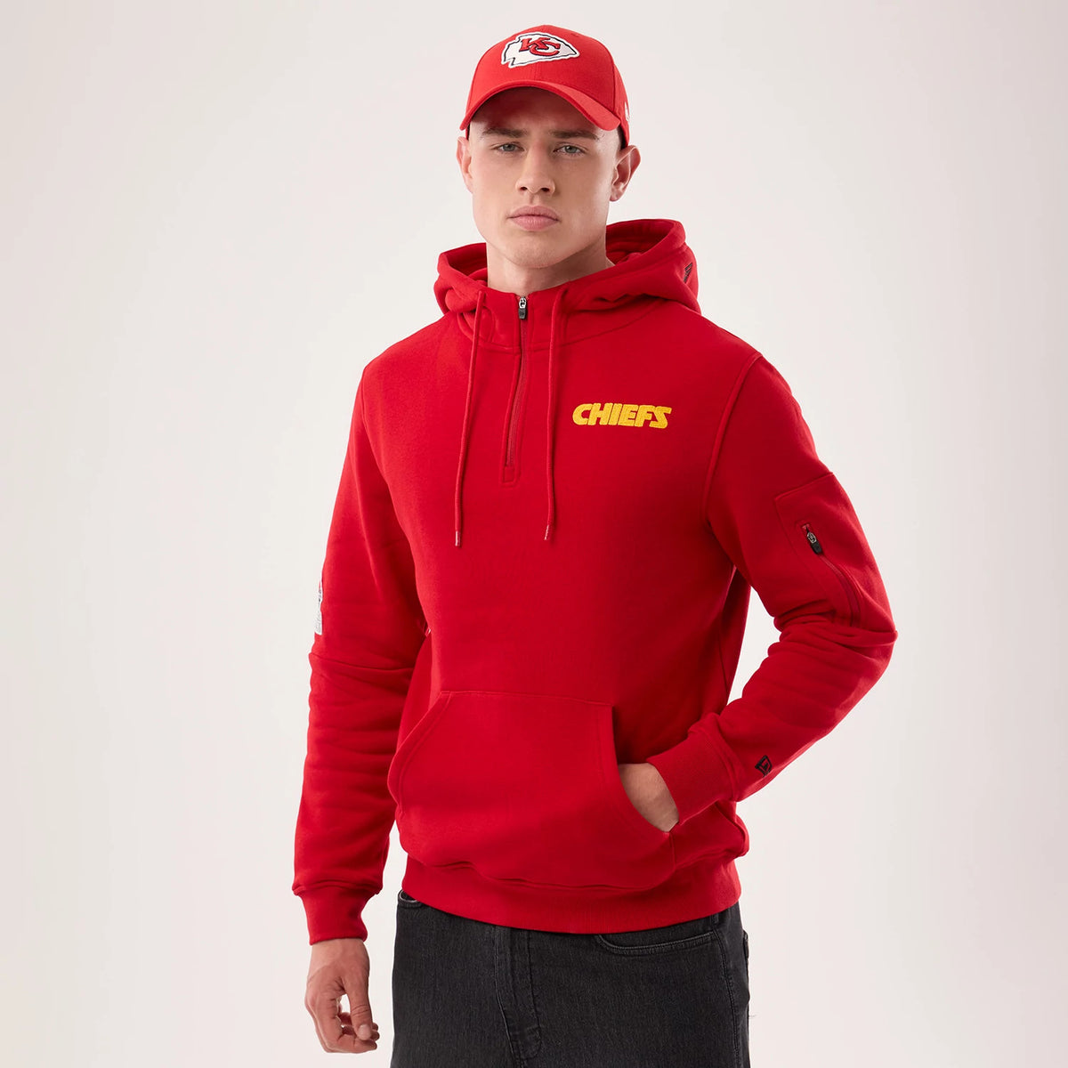 New Era Kansas City Chiefs NFL Sideline 2026 Red Pullover Hoodie, front view on-model showing the yellow Chiefs print on the left chest, a half-zip front, a zip pocket on the sleeve, and ribbed cuffs, paired with a matching red cap.