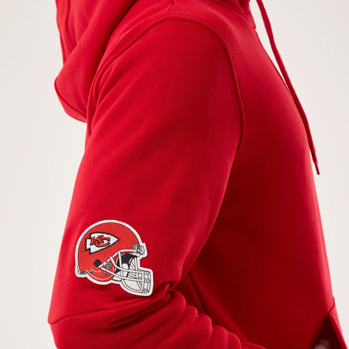 New Era Kansas City Chiefs NFL Sideline 2026 Red Pullover Hoodie, three-quarter view showing the vivid red fabric, a detailed embroidered Kansas City Chiefs helmet logo on the sleeve, and the hood partially behind the shoulder.