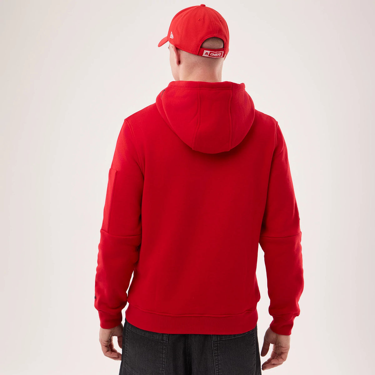 New Era Kansas City Chiefs NFL Sideline 2026 Red Pullover Hoodie, back view on a model wearing a matching cap, showing the plain red back with a relaxed hood draped over the shoulders and long sleeves without graphics.