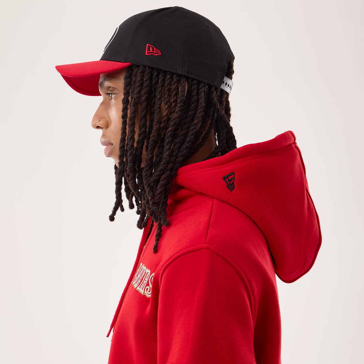 New Era San Francisco 49ers NFL Sideline 2026 Red Pullover Hoodie, side view on a model with a contrasting black and red cap, showcasing embroidered team lettering on the red hoodie and a small black team logo on the side of the hood.