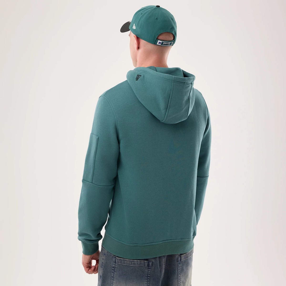 New Era Philadelphia Eagles NFL Sideline 2026 Dark Green Pullover Hoodie, back view on-model with a dark green hood, a debossed logo at the top, and ribbed cuffs, styled with a matching cap featuring an embroidered New Era logo on the side.