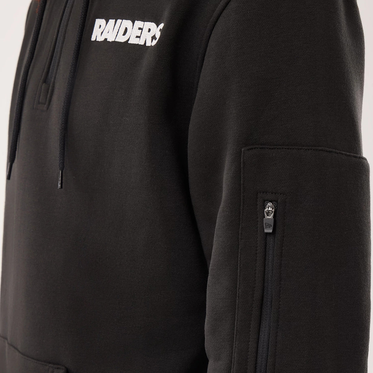 New Era Las Vegas Raiders NFL Sideline 2026 Black Pullover Hoodie, close-up side view showing the white embroidered Raiders lettering on the chest, an adjustable drawstring hood, and a small zipped pocket with a raised New Era logo on the sleeve.