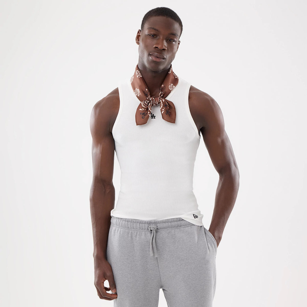 New Era Paisley Satin Brown Bandana, on-model view with a paisley pattern, tied around the neck in a neat bow, complementing a white tank top and grey joggers, showcasing versatile styling for a casual outfit.