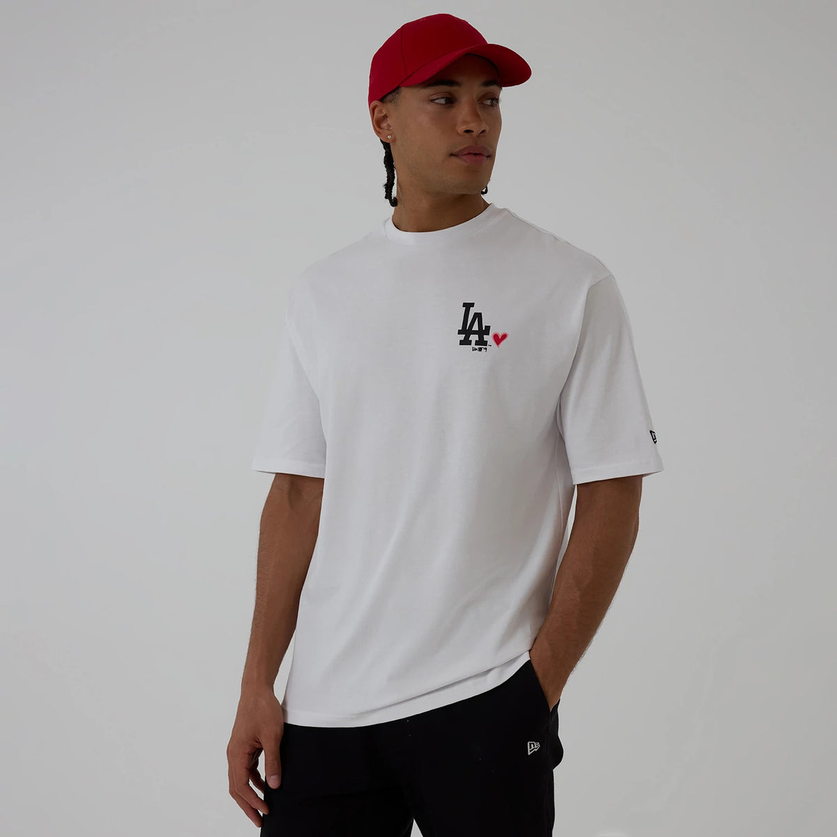 New Era LA Dodgers MLB Heart Graphic White T-Shirt, three-quarter view on a model wearing a red cap, showing the black LA logo with a red heart printed on the left chest and a small New Era embroidered logo on the left sleeve.