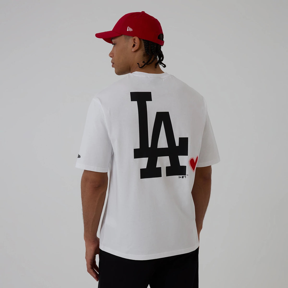 New Era LA Dodgers MLB Heart Graphic White T-Shirt, back view on a model wearing a red cap, showing a large black printed LA graphic centred on the white fabric with a small red heart graphic placed nearby on the lower right side.
